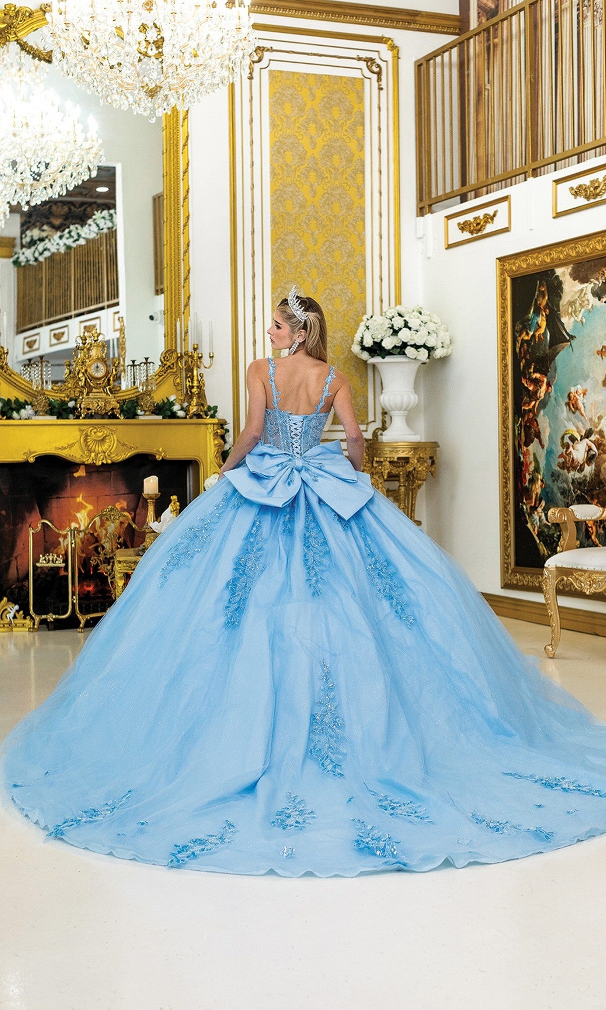 A woman wearing the Quinceanera Dress 5046 By Dancing Queen—a light blue ball gown with a corset bodice and large bow—stands in an ornate, gold-accented room with a chandelier, fireplace, and classical artwork.